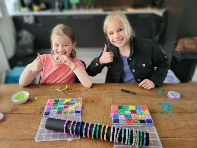 Twee meisjes met opgestoken rechterduim aan een houten tafel, waarop twee sorteerdozen met kraaltjes in allerlei kleuren liggen. Op de opengeklapte deksels van de sorteerdozen ligt een zwarte rol met daaromheen ongeveer vijftien armbandjes. Het meisje links is de zevenjarige Sara met donkerblond haar en een roze T-shirt, naast haar de hoogblonde elfjarige Isa met een paars shirtje en daarover een zwart jasje met opvallende lichte knopen.